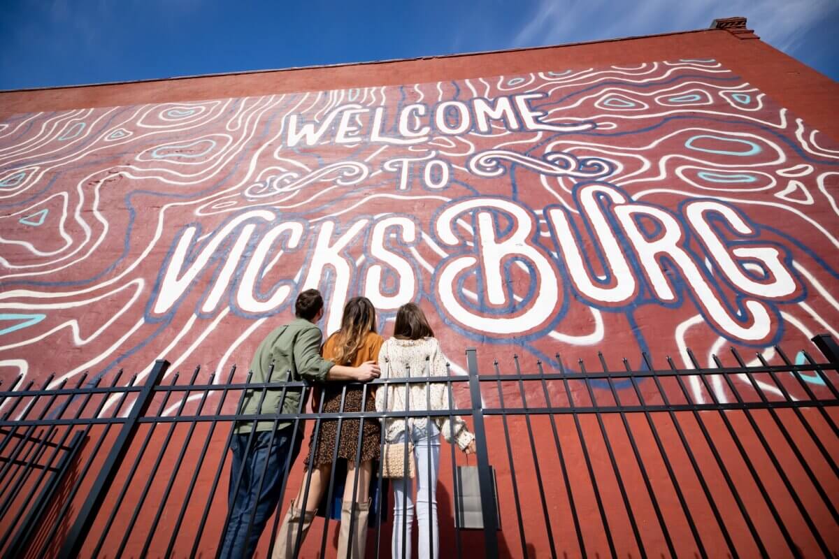 Three people stand with arms around each other, facing a large mural on a red brick wall that says "Welcome to Vicksburg," highlighting this Mississippi city as an art destination.