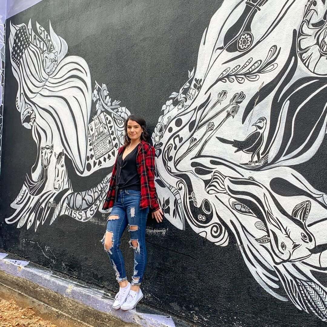 Why Vicksburg Might Be Mississippi's Most Unexpected Art Destination - 4 A woman in a red plaid shirt and ripped jeans stands in front of a black-and-white mural featuring abstract animal and nature designs, highlighting Vicksburg, Mississippi as an emerging art destination.