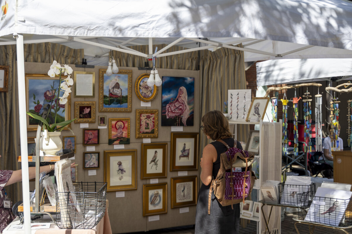Why Vicksburg Might Be Mississippi's Most Unexpected Art Destination - 9 A person with a backpack browses framed artwork inspired by the Vicksburg murals, displayed on the walls of an outdoor art market booth under a white canopy.