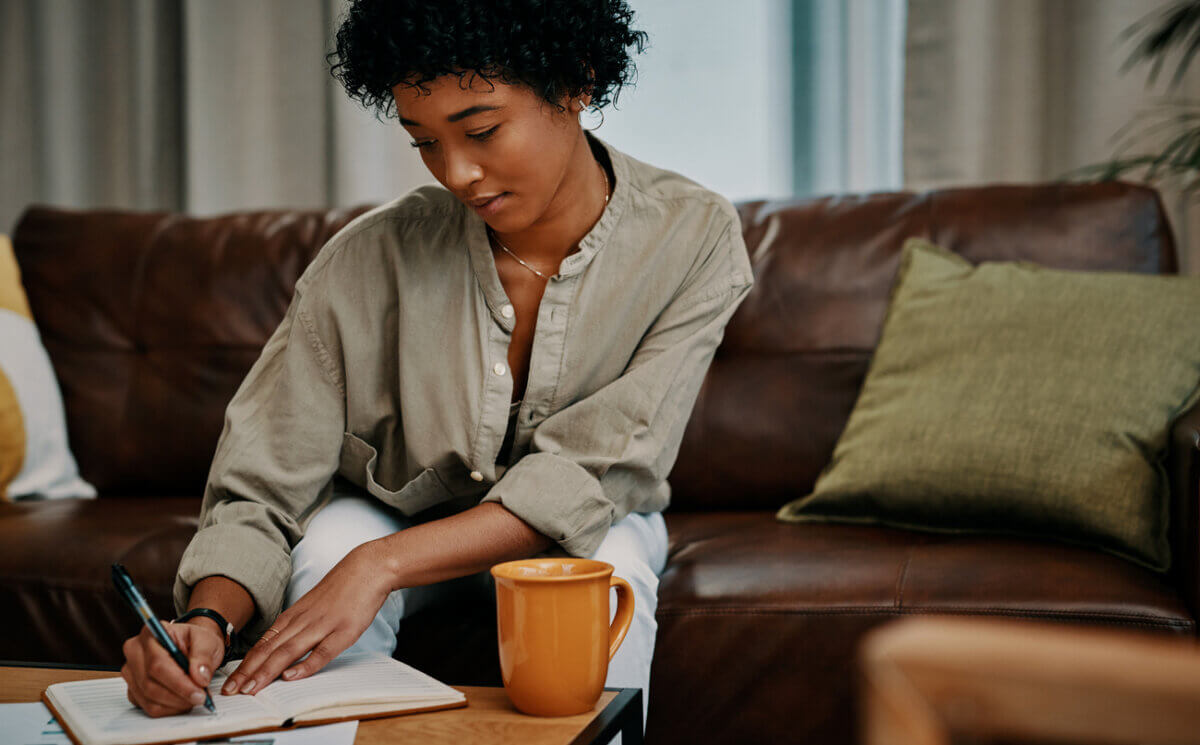 "Woo" Habits I Swear By for Financial Peace & Success - 3 A person sits on a couch, writing in a notebook at a coffee table with an orange mug in front of them, reflecting on woo habits that contribute to their financial success.