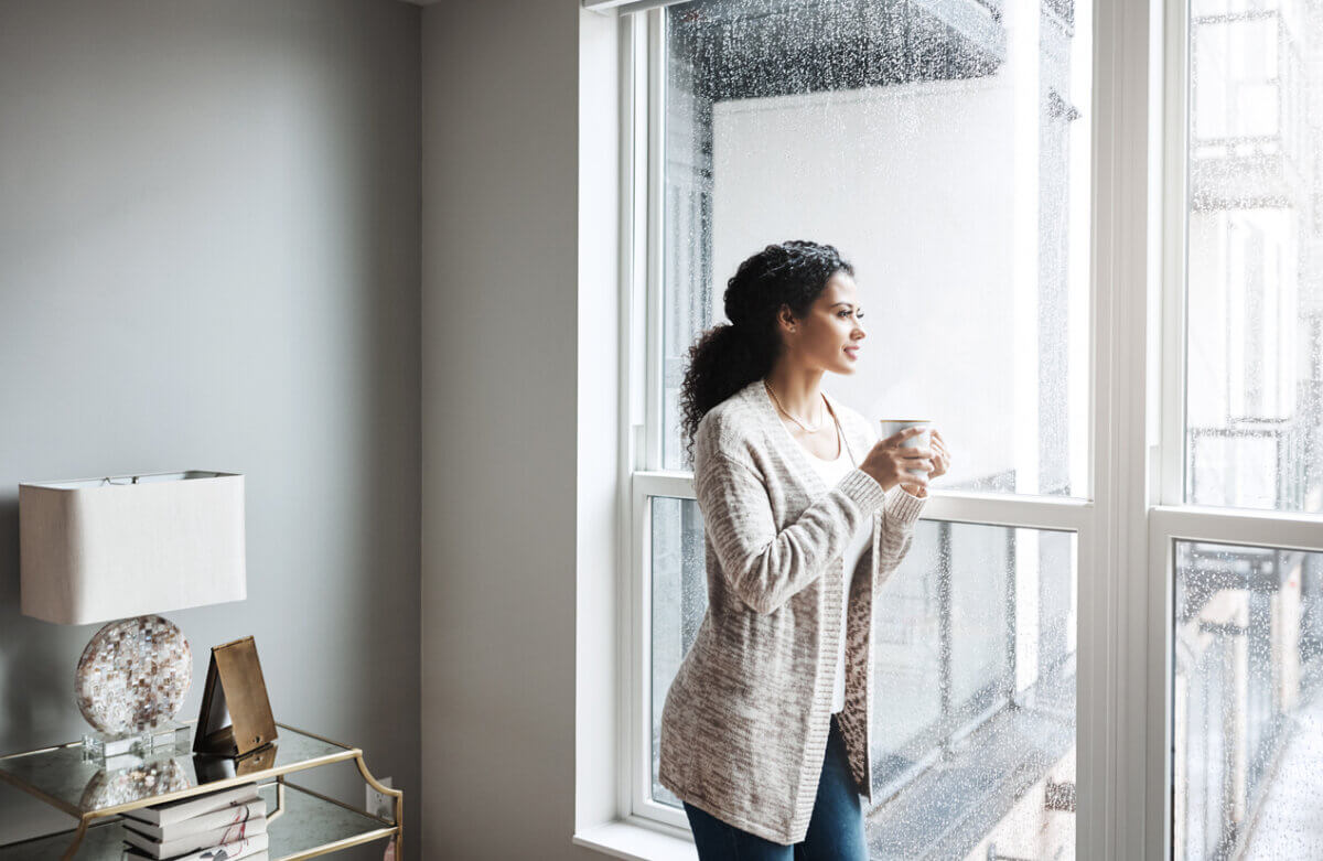 "Woo" Habits I Swear By for Financial Peace & Success - 2 Woman holding a mug stands by a large window, looking outside on a rainy day in a modern living room.