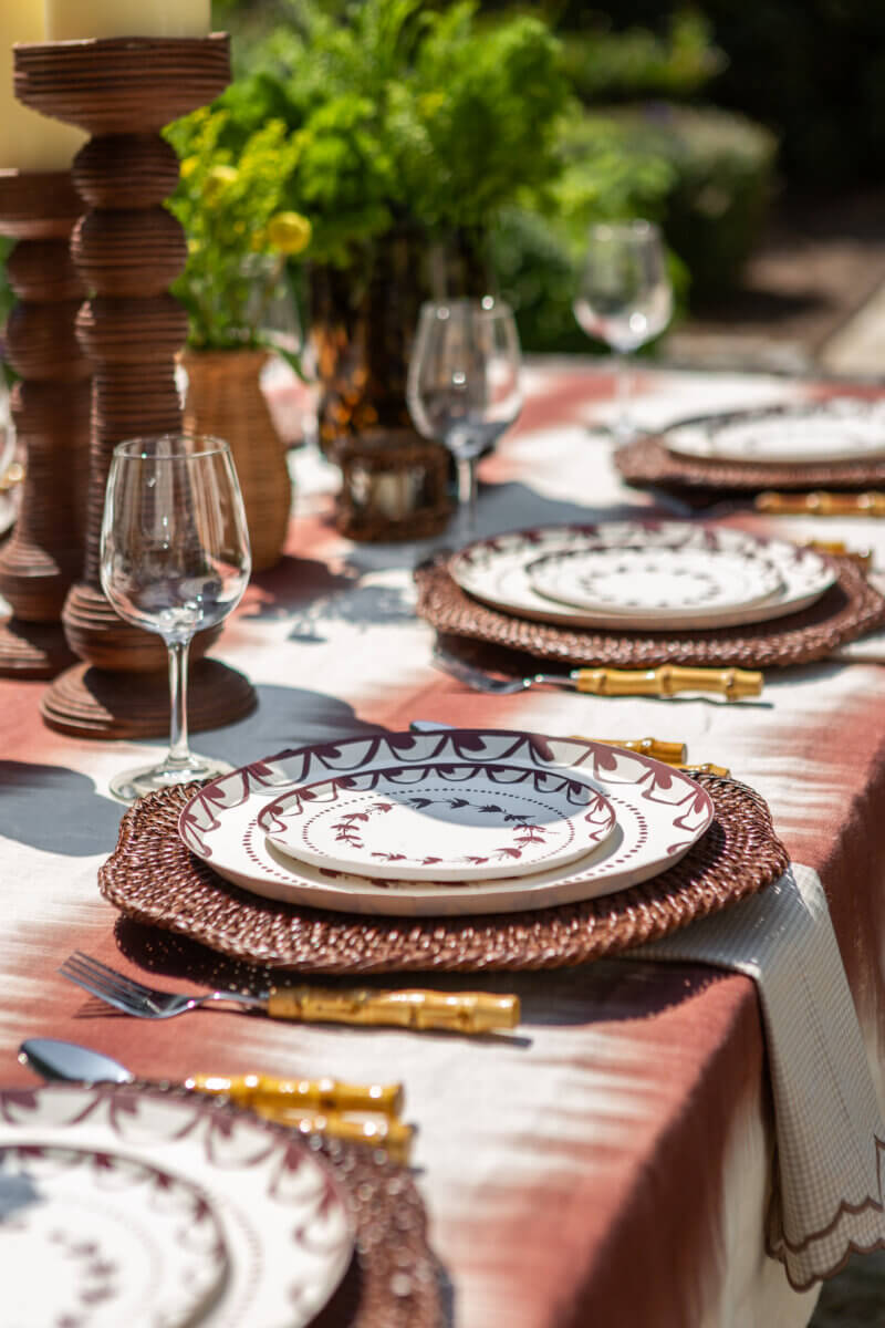 Table Two Studio's Luxe Paper Plates Will Change How You Entertain - 5 A table set for a meal outdoors with Table Two Studio luxury paper plates, woven placemats, glassware, gold utensils, and decorative candles and greenery arranged at the center.