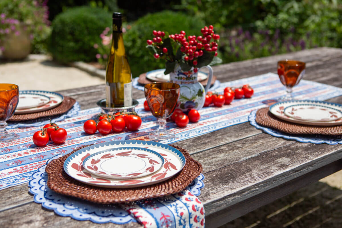 Table Two Studio's Luxe Paper Plates Will Change How You Entertain - 10 A rustic wooden table set for four with Table Two Studio luxury paper plates, amber glasses, a bottle of wine, tomatoes, and a floral centerpiece, awaits guests outdoors in a charming garden.