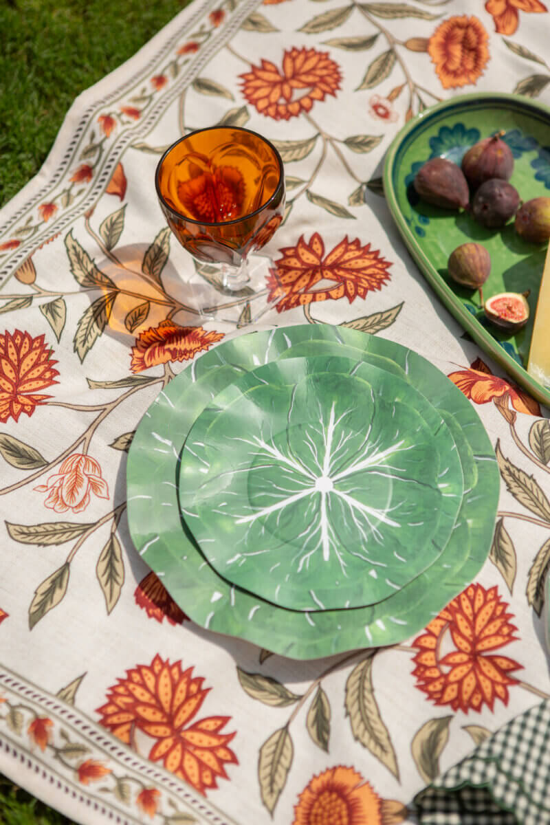 Table Two Studio's Luxe Paper Plates Will Change How You Entertain - 7 A picnic setup with Table Two Studio luxury paper plates featuring green leaf patterns, an amber glass, and a green tray with figs and cheese, all arranged on a floral-patterned cloth laid out on the grass.