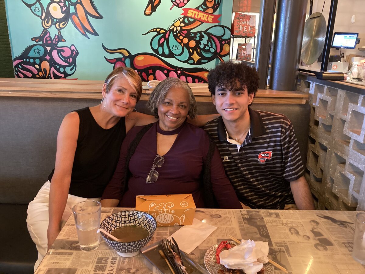 Meet the Teacher Generations of Nashville Kids Call “Ms. Juanita” - 4 Three people sit closely together and smile at a restaurant booth with colorful wall art in the background. Food and drinks are on the table.