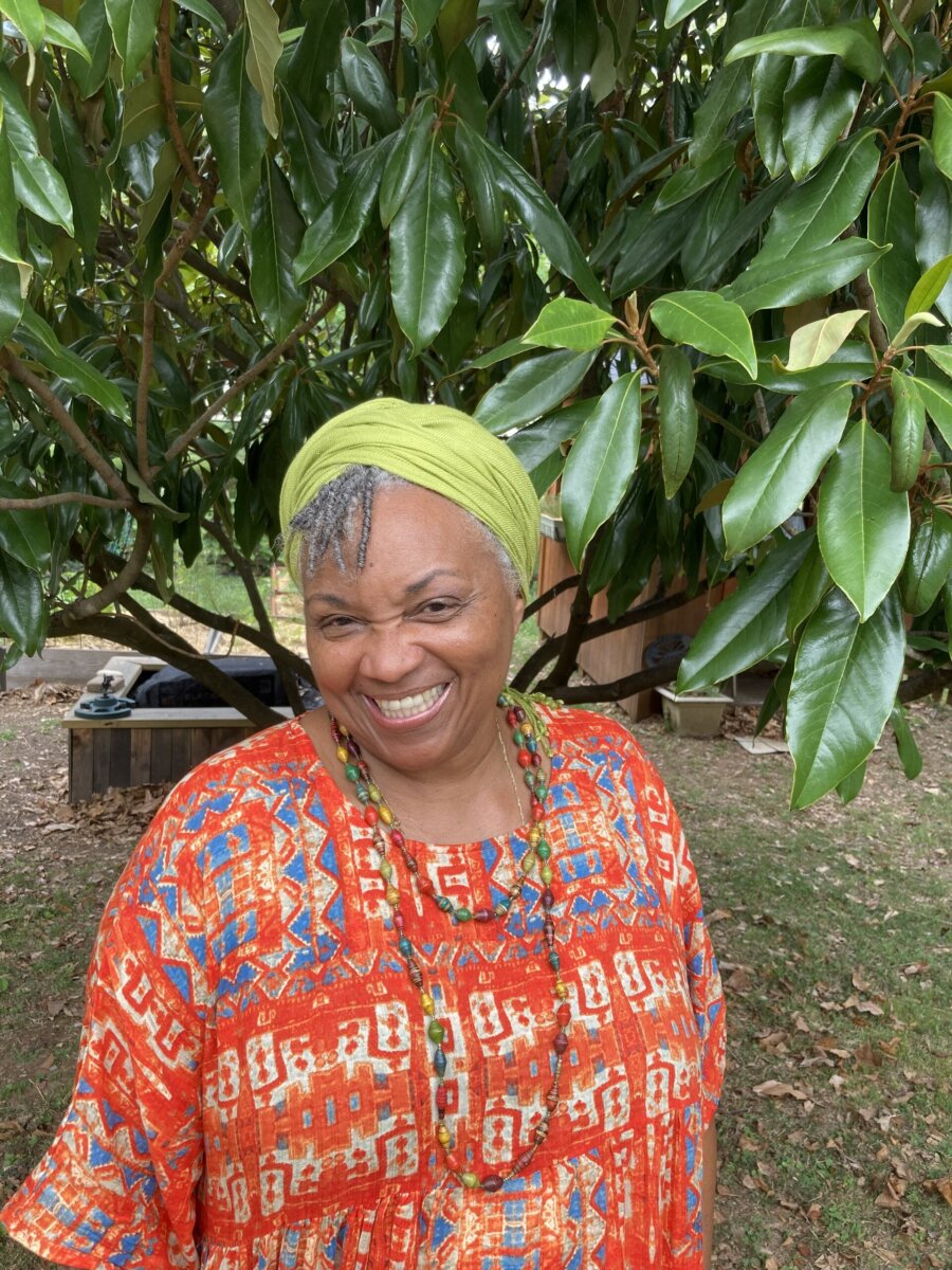 Meet the Teacher Generations of Nashville Kids Call “Ms. Juanita” - 3 Ms. Juanita, wearing a green headwrap and a red patterned top, stands outdoors in front of a tree with broad green leaves, radiating warmth.