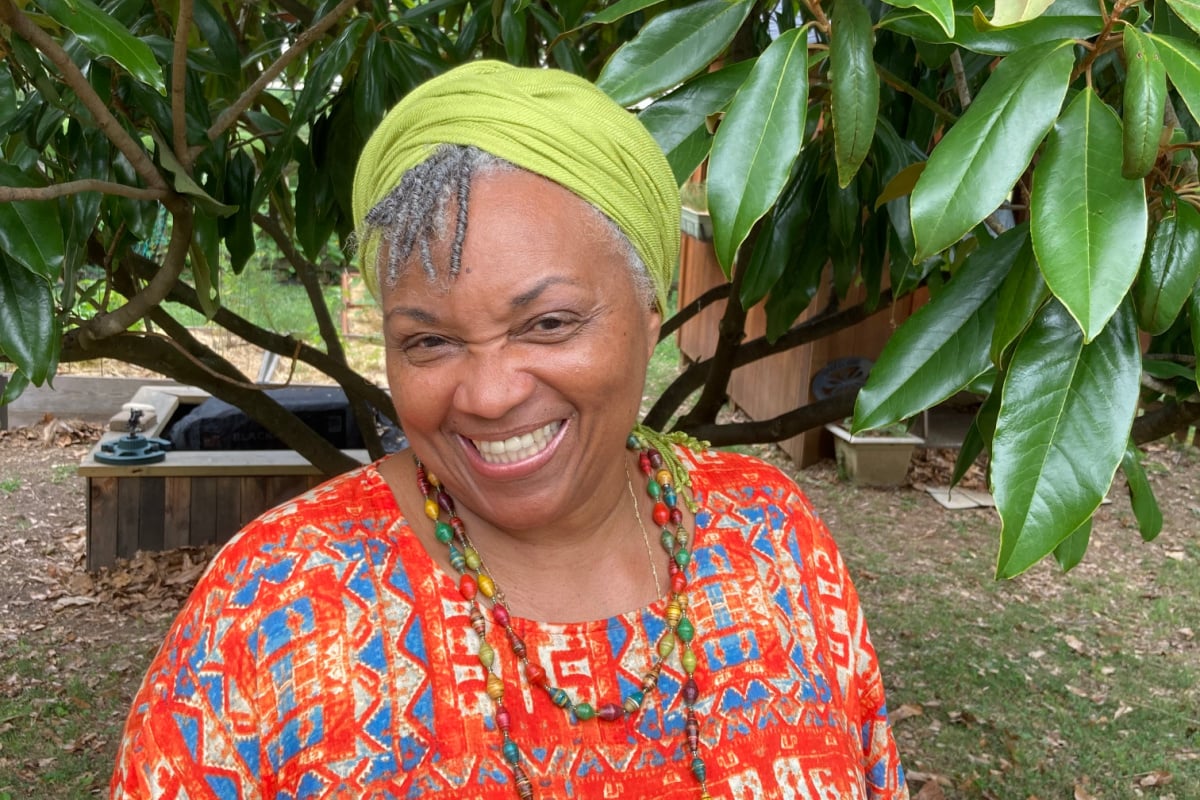 Ms. Juanita, wearing a green headscarf and colorful patterned top, smiles while standing in front of leafy green branches outdoors.