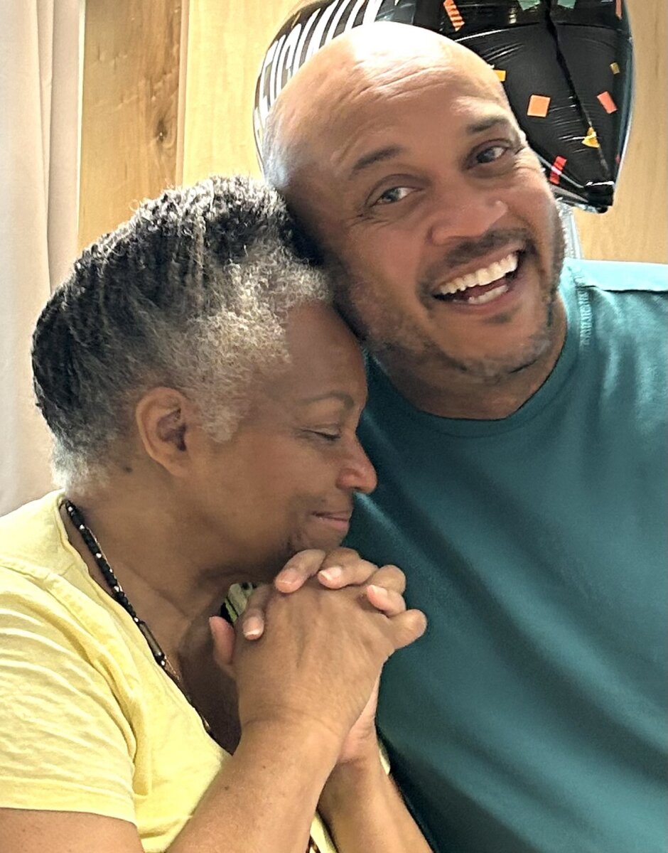 Meet the Teacher Generations of Nashville Kids Call “Ms. Juanita” - 2 Two people are standing close together indoors; one is smiling at the camera while the other leans on their shoulder, holding hands and appearing emotional.