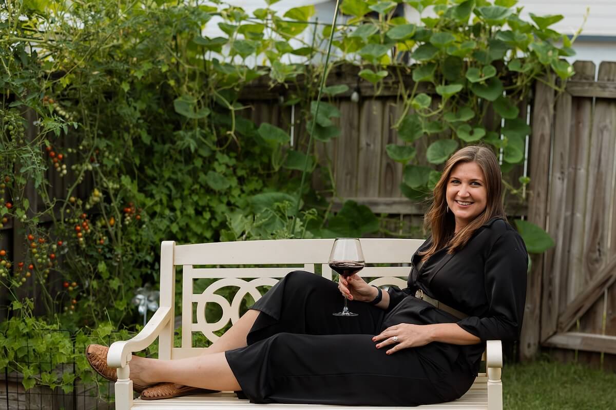 Lauren Maranto sits on a garden bench in a black outfit, her FACE illuminated by a smile as she holds a glass of red wine, surrounded by green plants and a wooden fence.