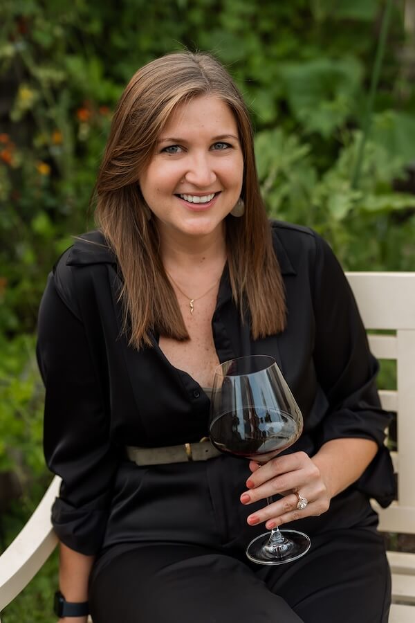 Meet the Sommelier Behind Nashville's Newest Wine Bar + Bodega - 2 Lauren Maranto sits on a white bench outdoors in a black outfit, holding a glass of red wine and smiling at the camera, framed by green foliage in the background.