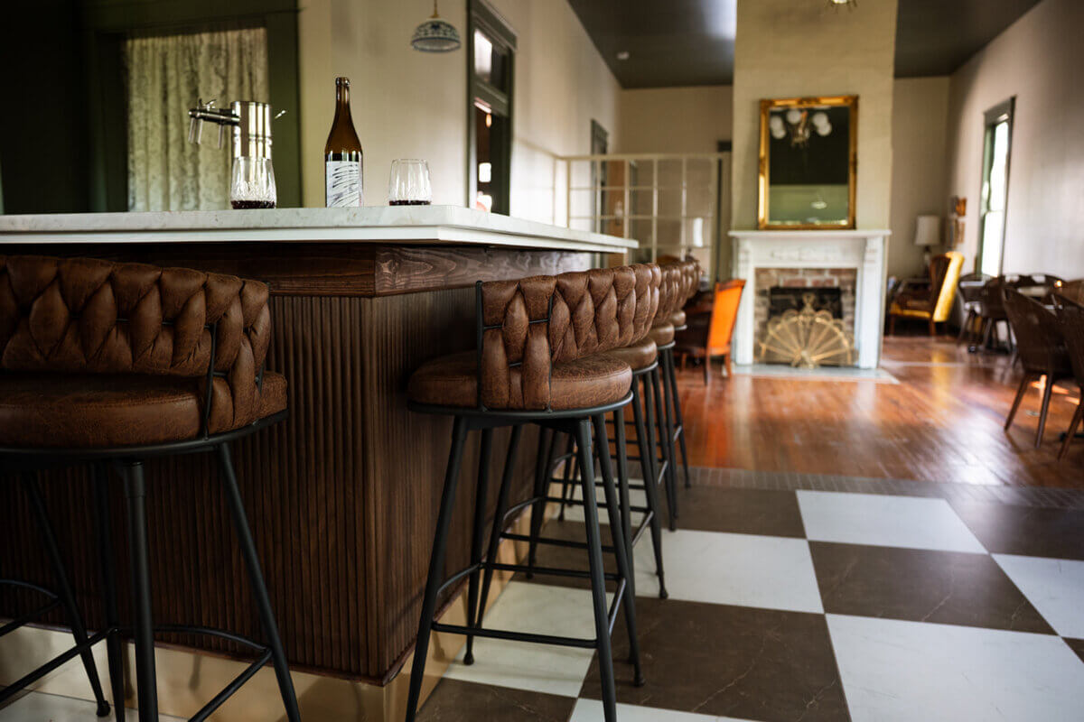 Meet the Sommelier Behind Nashville's Newest Wine Bar + Bodega - 4 A bar area with tufted leather stools, a marble countertop, a bottle and glasses, and a view into a spacious Lauren Maranto-designed lounge with a fireplace and wooden floors.