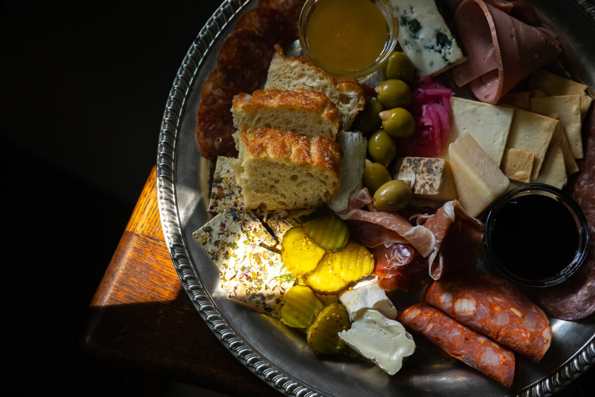 Meet the Sommelier Behind Nashville's Newest Wine Bar + Bodega - 3 A silver tray by Lauren Maranto holds assorted meats, cheeses, pickles, olives, bread, crackers, and two small containers of dipping sauce, with sunlight highlighting part of the arrangement.