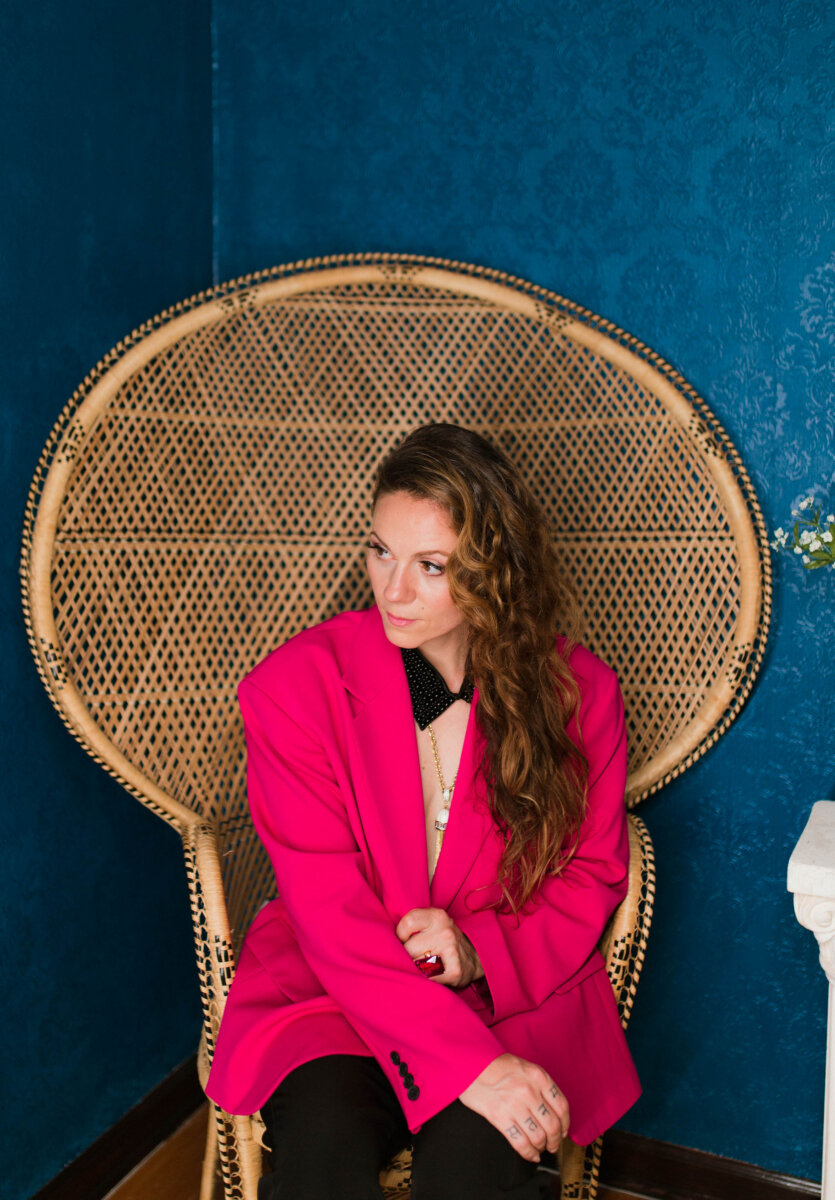 Ever Heard of a “Book Doula?" Meet Rea Frey - 6 Rea Frey, with long wavy hair, sits on a wicker chair against a blue wall, wearing a bright pink blazer over a black outfit and looking off to the side.
