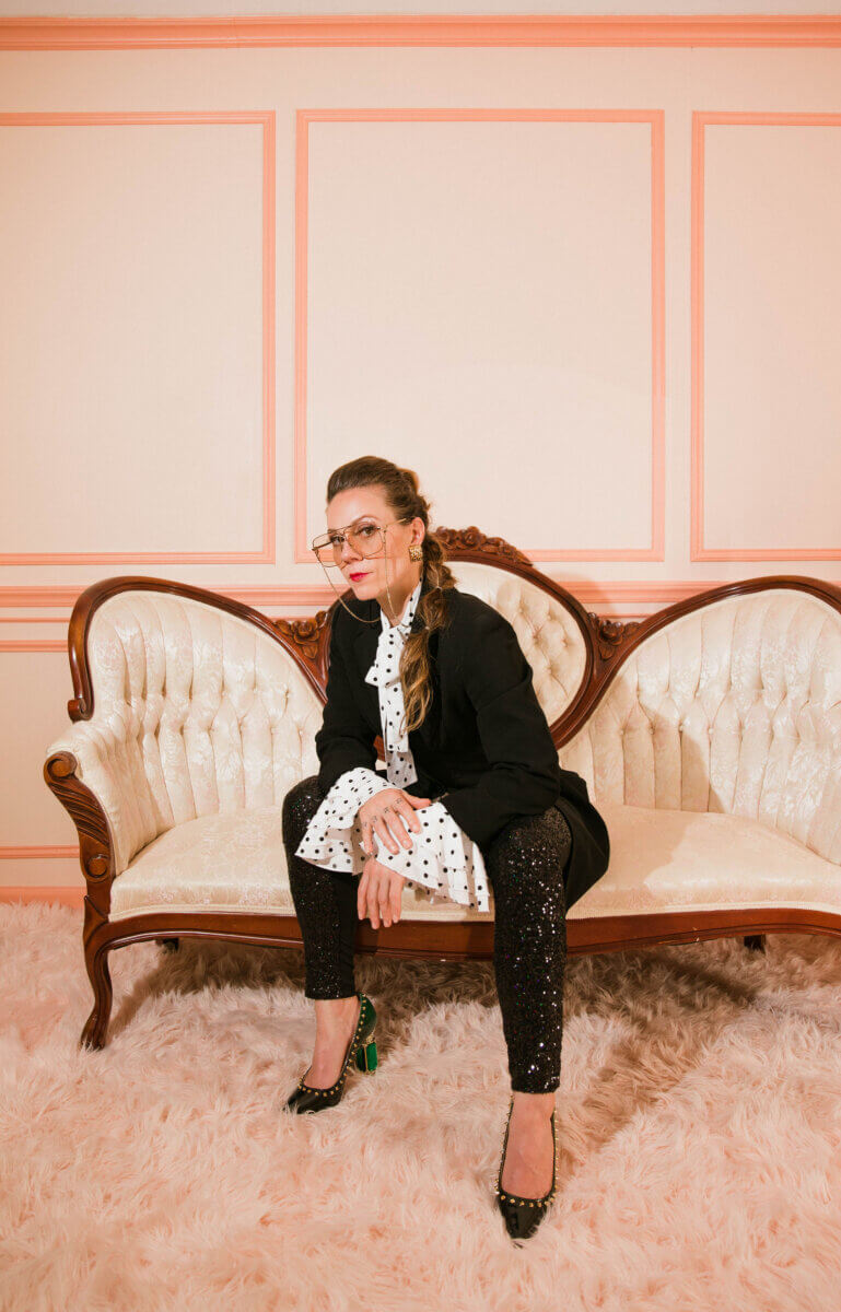 Ever Heard of a “Book Doula?" Meet Rea Frey - 4 Rea Frey, wearing a black blazer, polka dot blouse, sequined pants, and heels, sits confidently on a vintage cream sofa in a pink-walled room with glasses and a shag rug.