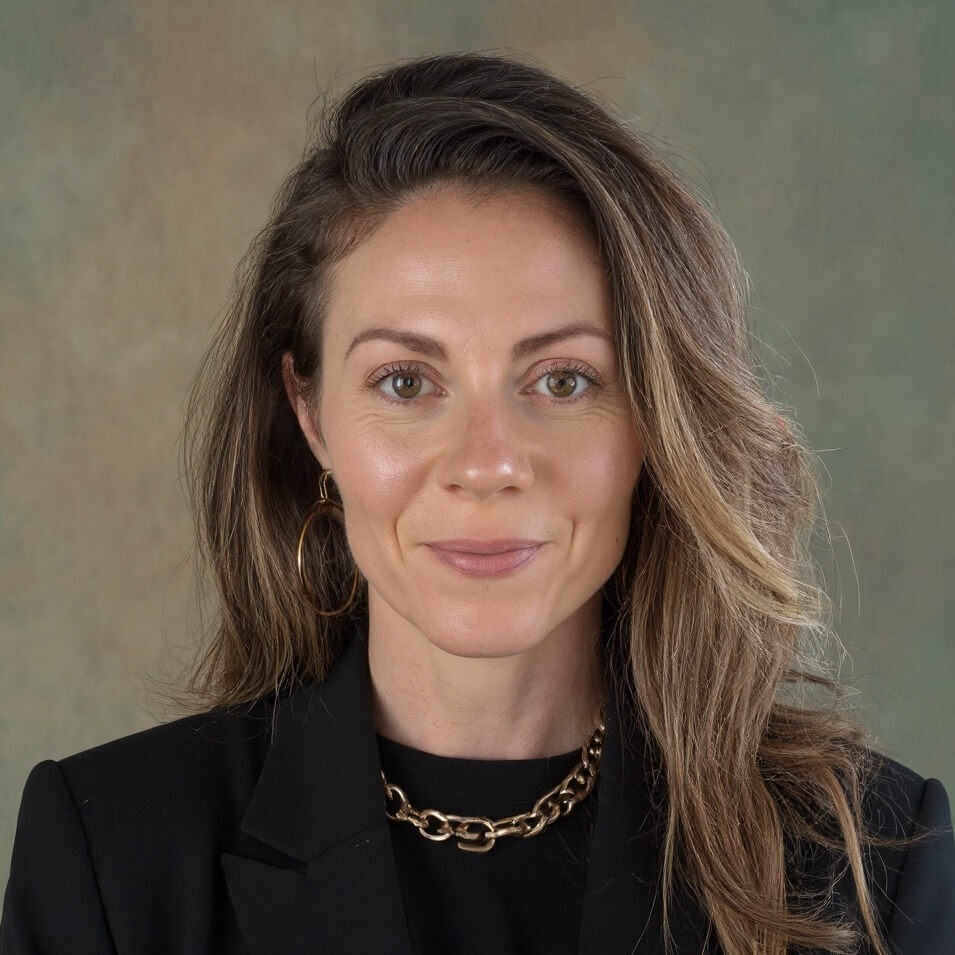 Ever Heard of a “Book Doula?" Meet Rea Frey - 2 Rea Frey, a woman with long brown hair, wears a black blazer, gold chain necklace, and hoop earring as she poses confidently in front of a neutral background.