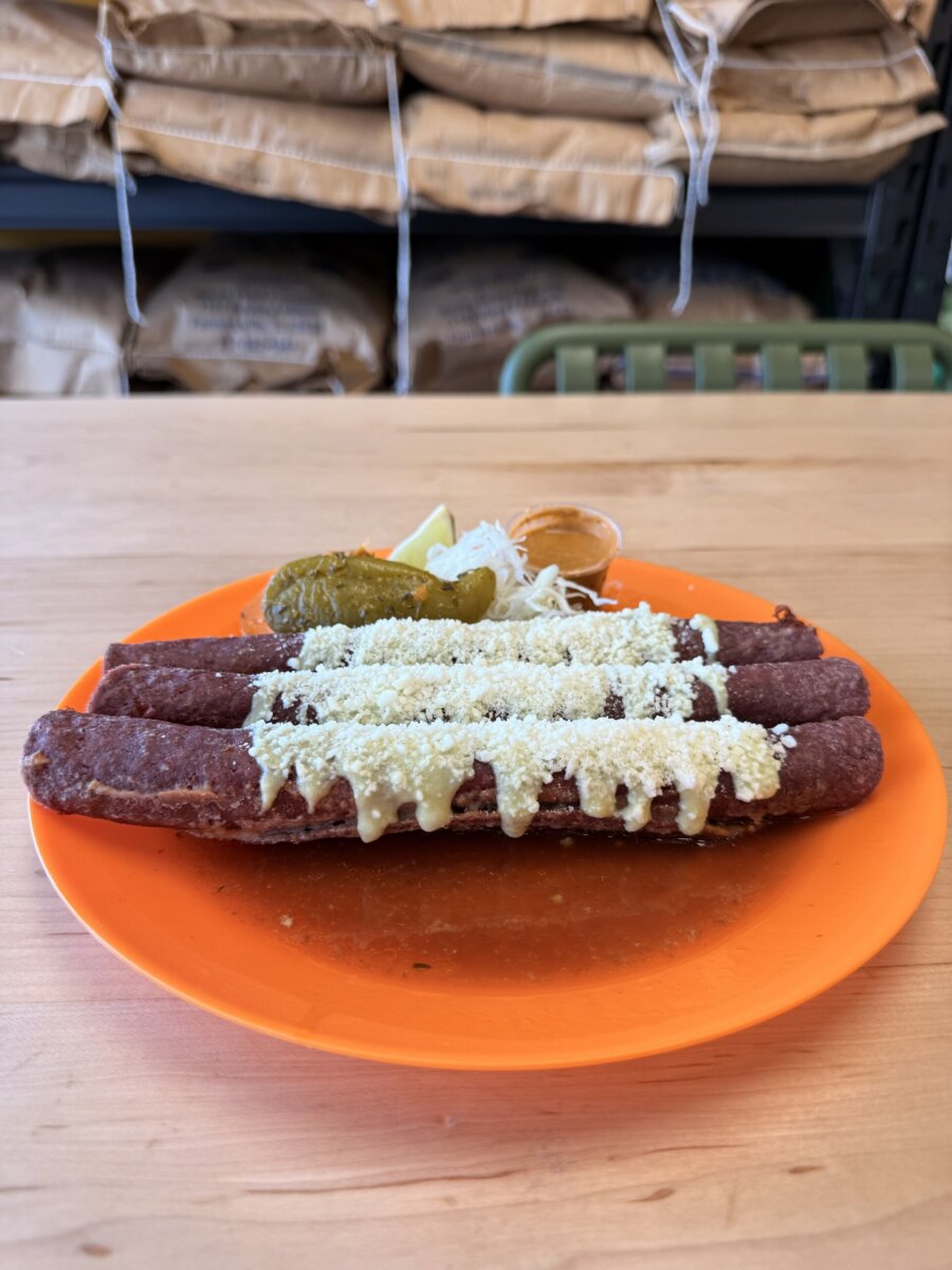7 New Nashville Restaurants to Try This Month - 4 Three sausages topped with cheese sauce and grated cheese on an orange plate, served with pickles, shredded cabbage, a lime wedge, and a small cup of sauce—a must-try at new restaurants in Nashville by May 2026.