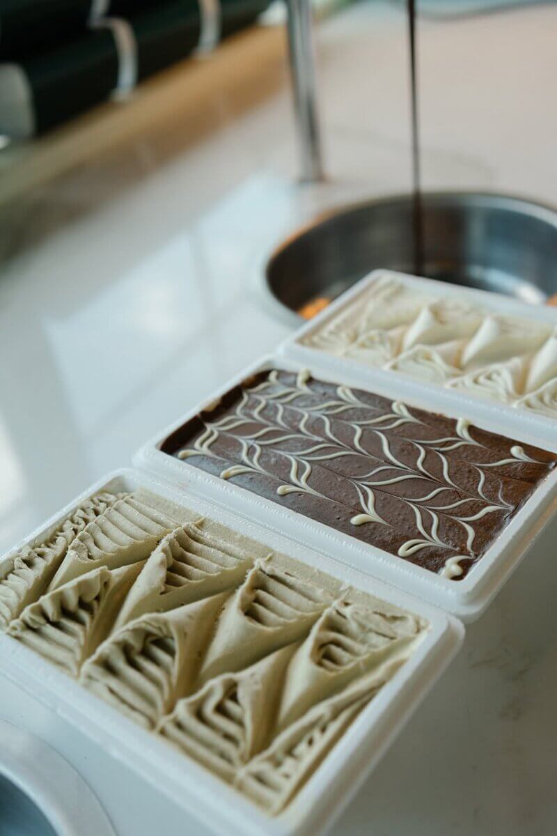 Three containers of gelato, two with light cream flavors and one with chocolate in the middle, are displayed on a counter near a metal sink.