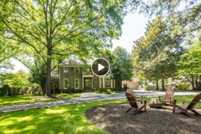 A two-story brick house with large windows sits behind a circular driveway, surrounded by tall trees and a yard with Adirondack chairs around a fire pit.