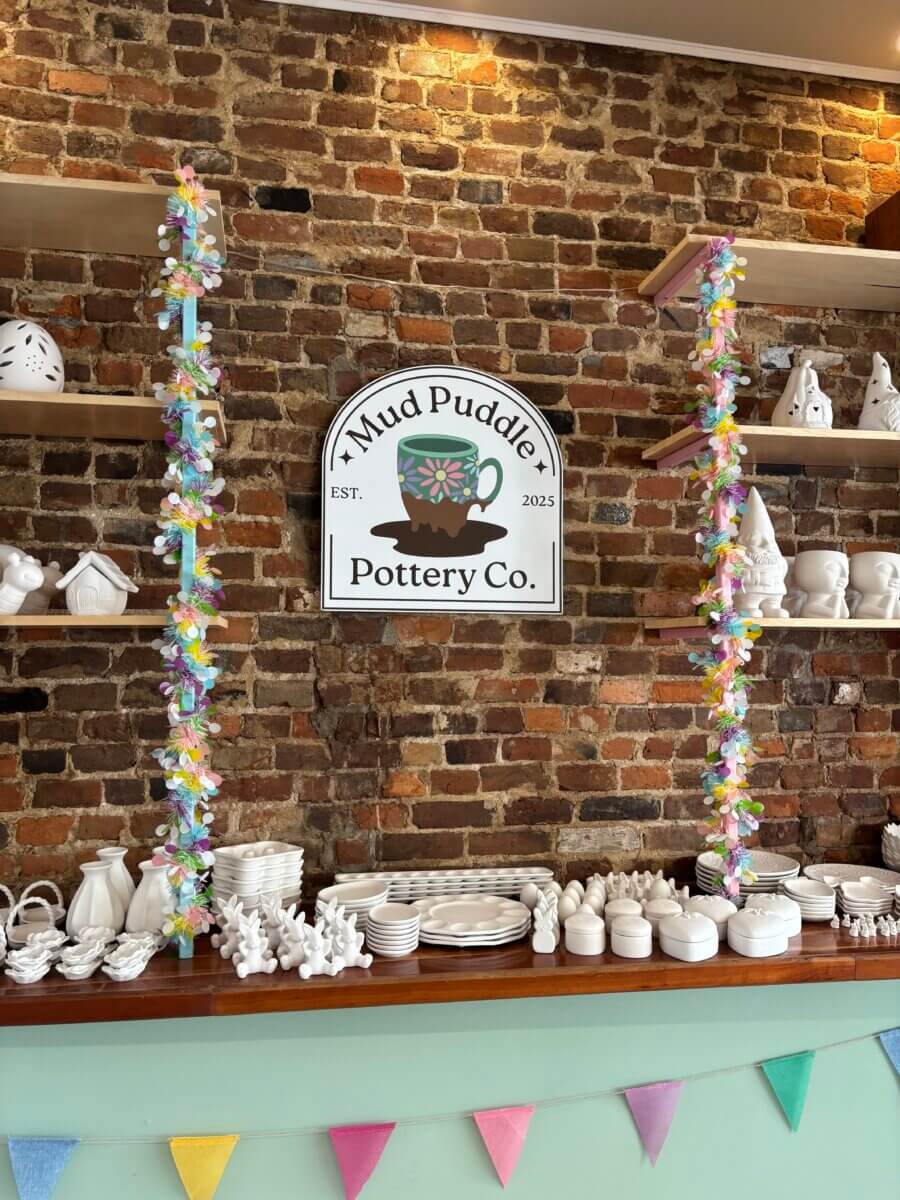Need a Change of Scenery? Try These Summer Weekend Trips From Nashville - 8 Shelves with unpainted pottery pieces and stacked plates against a brick wall, featuring a "Mud Puddle Pottery Co." sign and colorful garlands—perfect for creative weekend trips from Nashville.