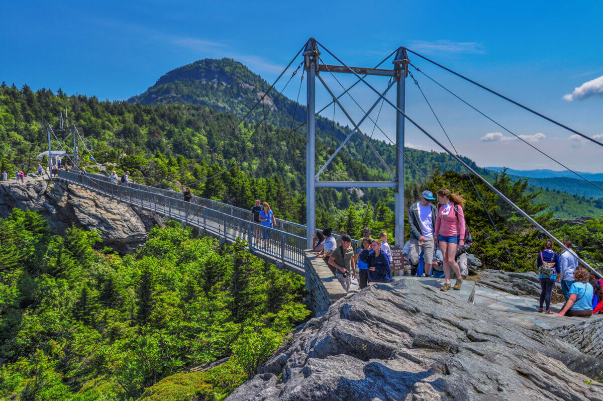 4 Summer Weekend Trips from Atlanta - 6 People walk across a suspension bridge and gather on rocky terrain surrounded by trees and mountains under a clear blue sky—an ideal scene for Summer Weekend Trips from Atlanta.