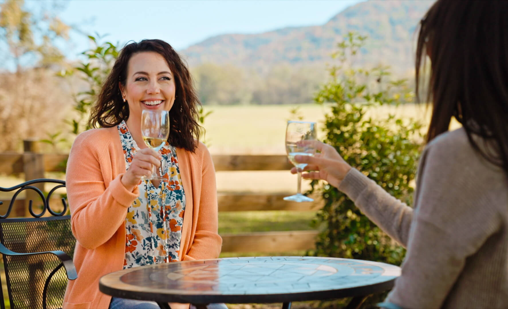 Two women sit at an outdoor table smiling and clinking wine glasses against a backdrop of mountains and greenery—capturing the perfect moment during a summer weekend trip.
