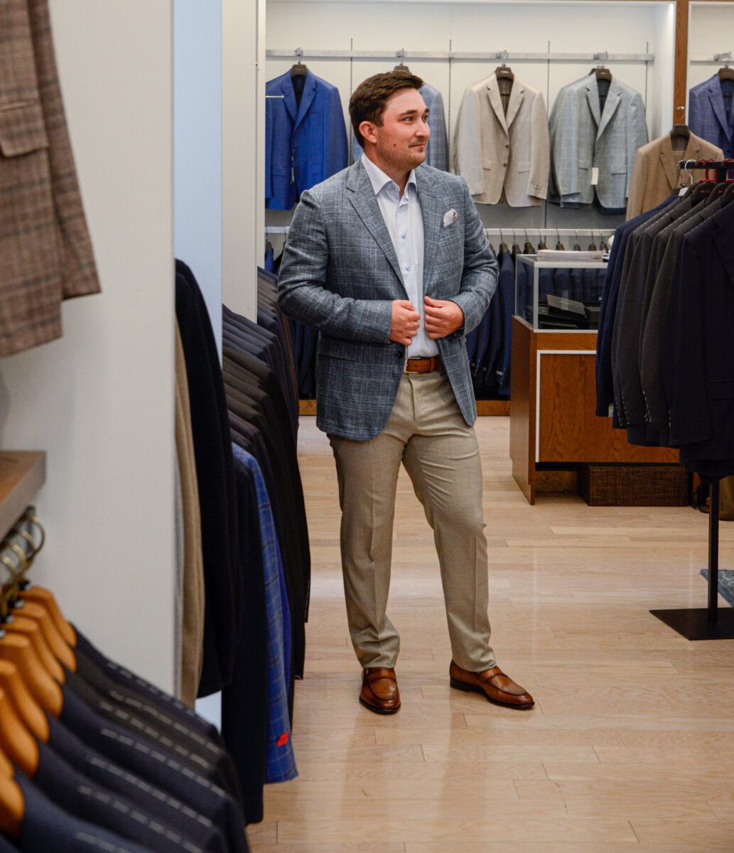 The Event-Season Trends Nashville Boutiques Are Loving Right Now - 13 A man in a suit jacket, dress shirt, and dress pants stands all dressed up in a men's clothing store, surrounded by hanging jackets and blazers.