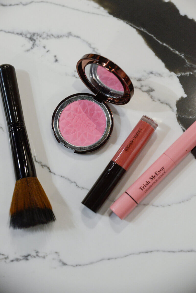The Event-Season Trends Nashville Boutiques Are Loving Right Now - 16 A makeup brush, pink blush compact, pink lip crayon, and a tube of lip gloss arranged on a marble surface.