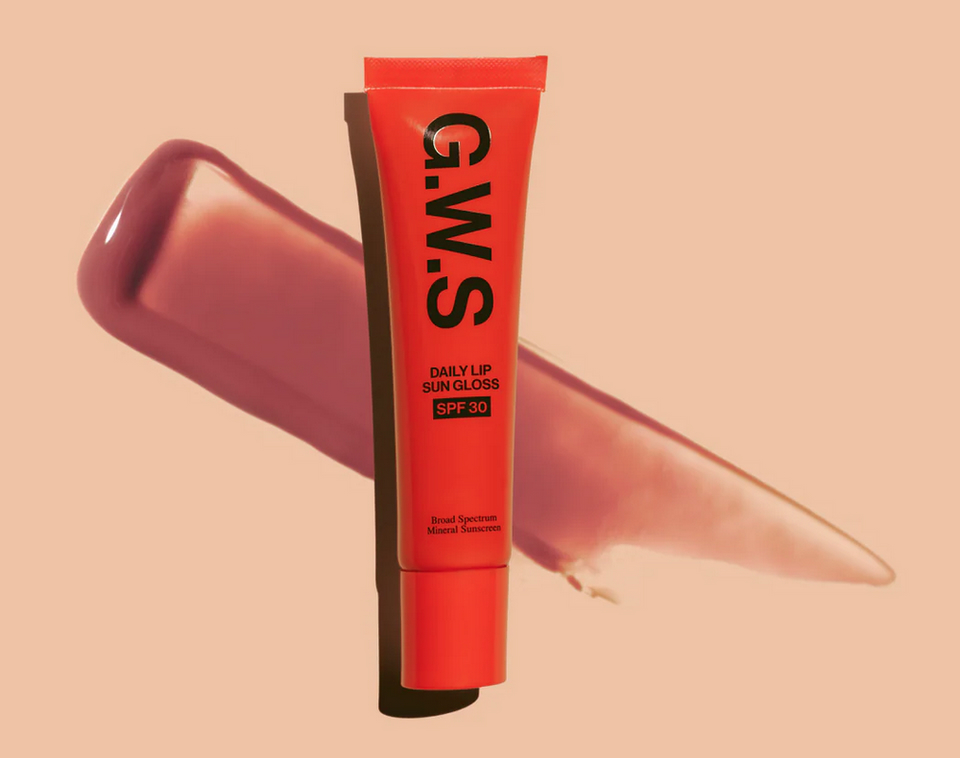 A tube of G.W.S Daily Lip Sun Gloss SPF 30 stands upright in front of a smear of pink gloss on a peach background, making it a must-have for summer beauty and heatproof makeup routines.