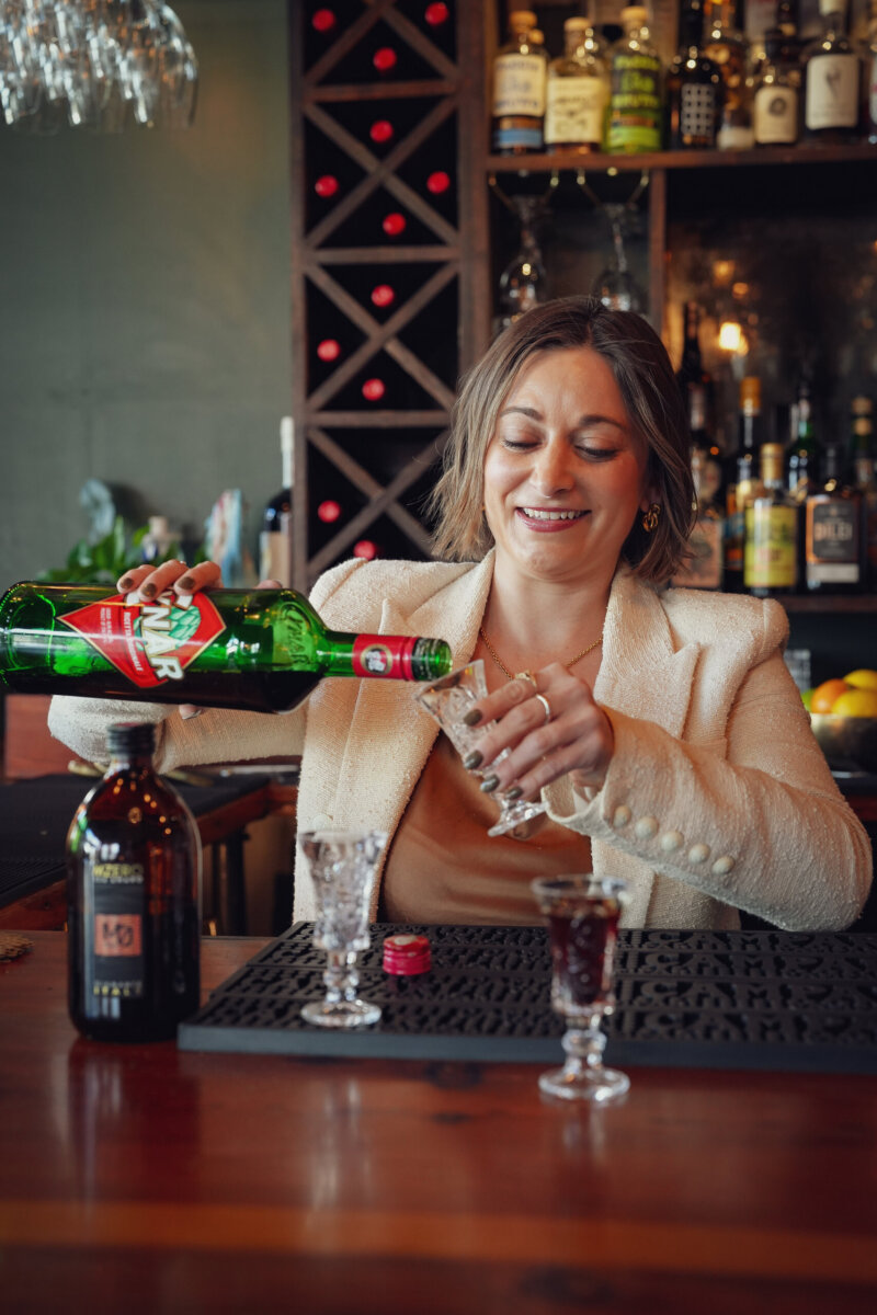 From Giving Shots to Serving Them: Meet Chattanooga's Cocktail Queen - 5 A woman behind the bar pours a green bottle of Cynar into a glass, with bottles of alcohol and wine on shelves in the background next to the beverage menu.