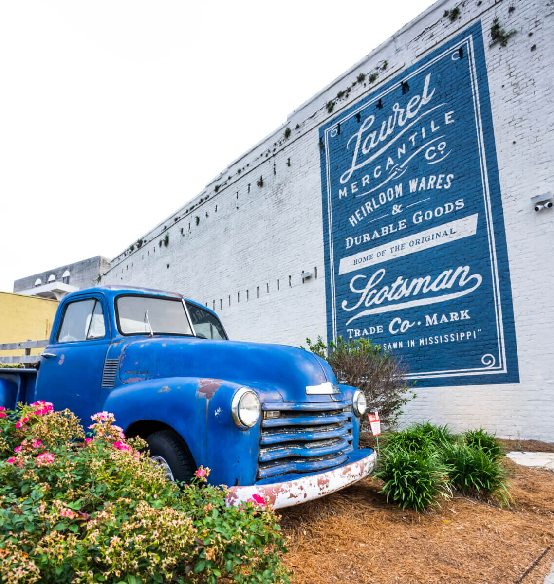 The Best Summer Weekend Getaways in the South - 6 A vintage blue pickup truck is parked in front of a white brick wall featuring a large Laurel Mercantile Co. advertisement mural—perfect inspiration for summer weekend trips in the South.