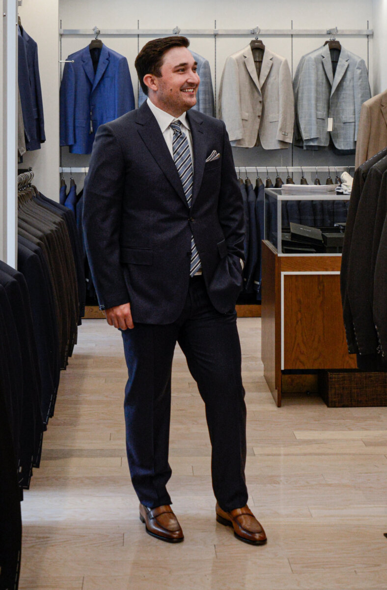 The Event-Season Trends Nashville Boutiques Are Loving Right Now - 11 All dressed up in a dark suit, white shirt, and striped tie, a man stands smiling in a store aisle lined with men's suits and jackets.
