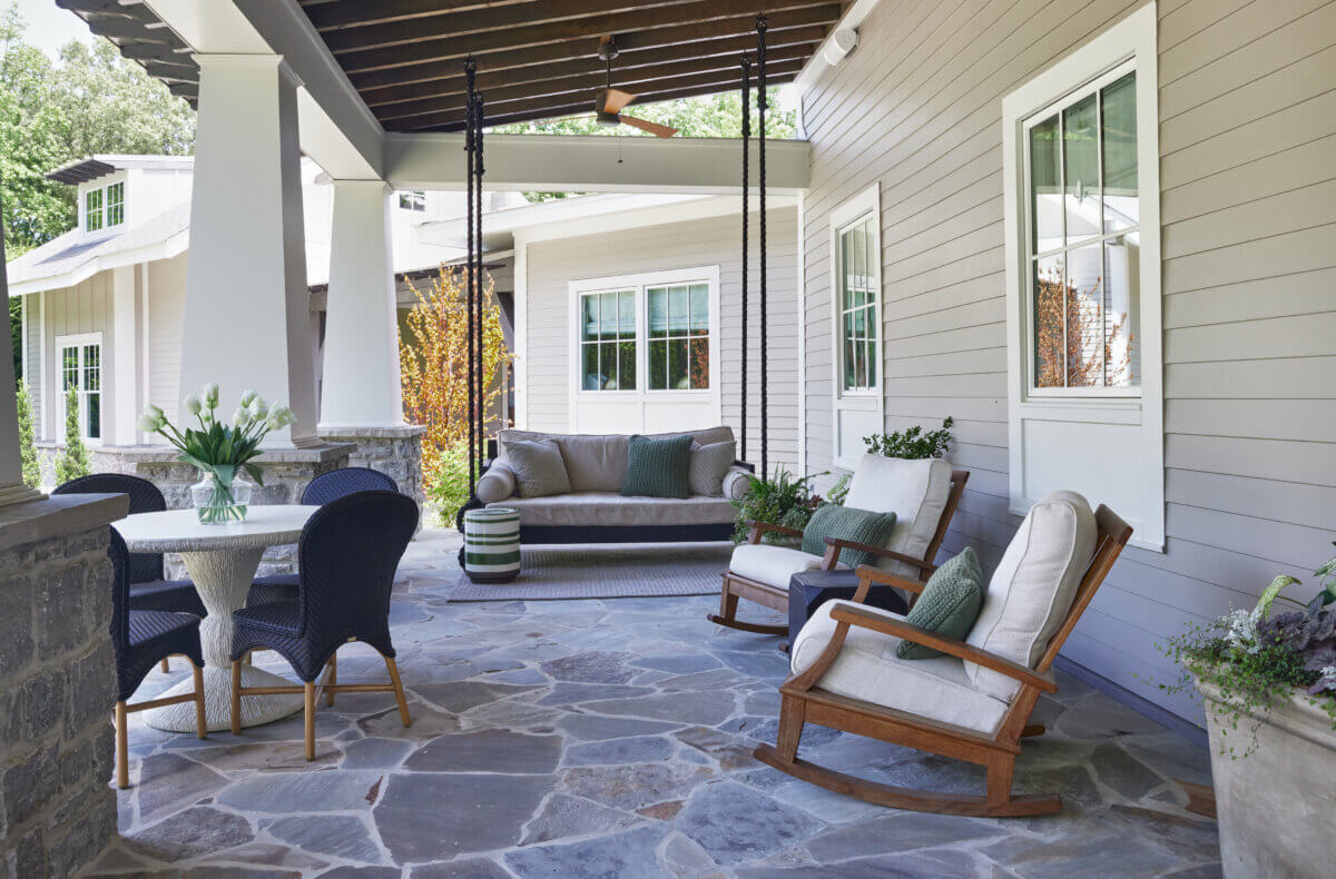 10 Designer Tips for Upgrading Your Outdoor Spaces - 9 Covered patio with stone flooring, cushioned swing sofa, rocking chairs, a round table with four chairs, potted plants, and a window on a light-colored house exterior.
