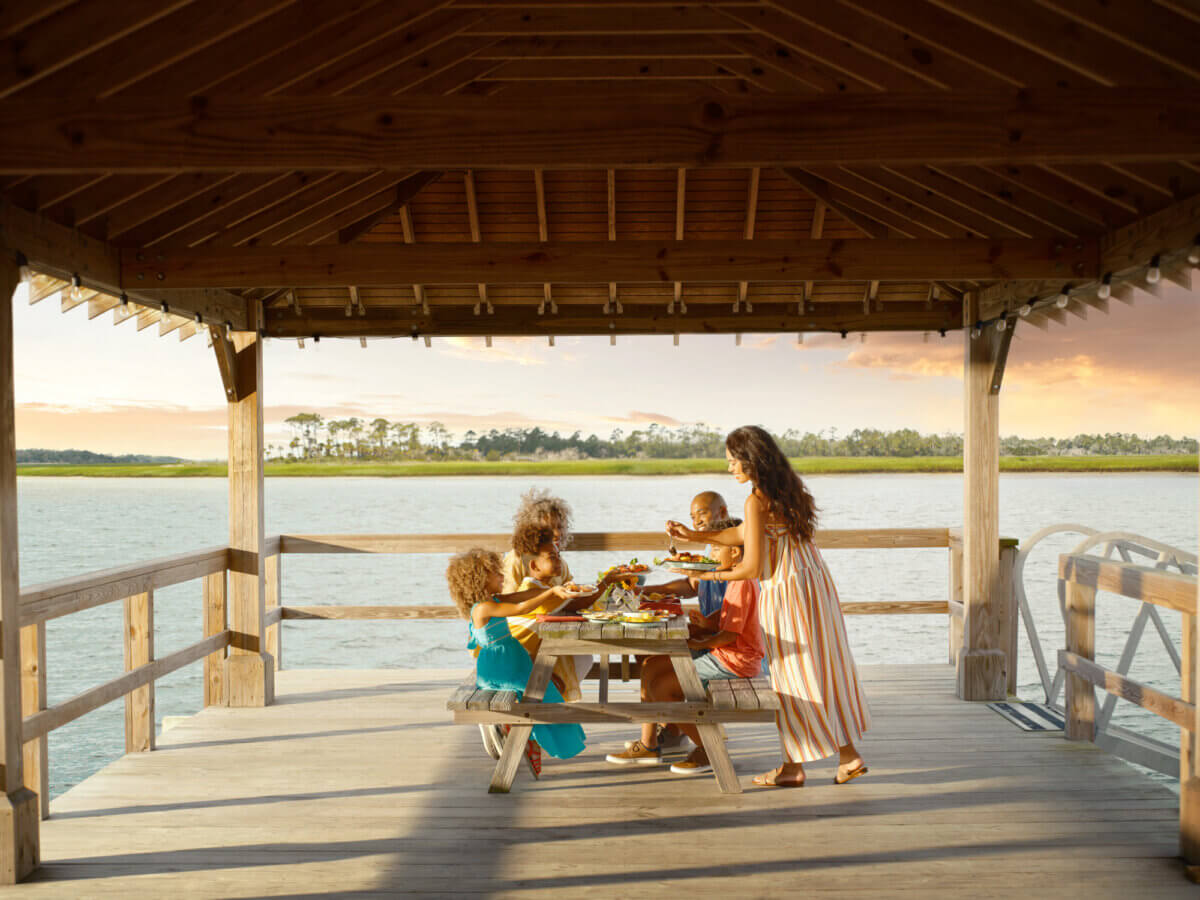 Four people share a meal at a wooden picnic table under a gazebo on a dock, overlooking the water during sunset—a perfect scene for summer weekend trips in the South.