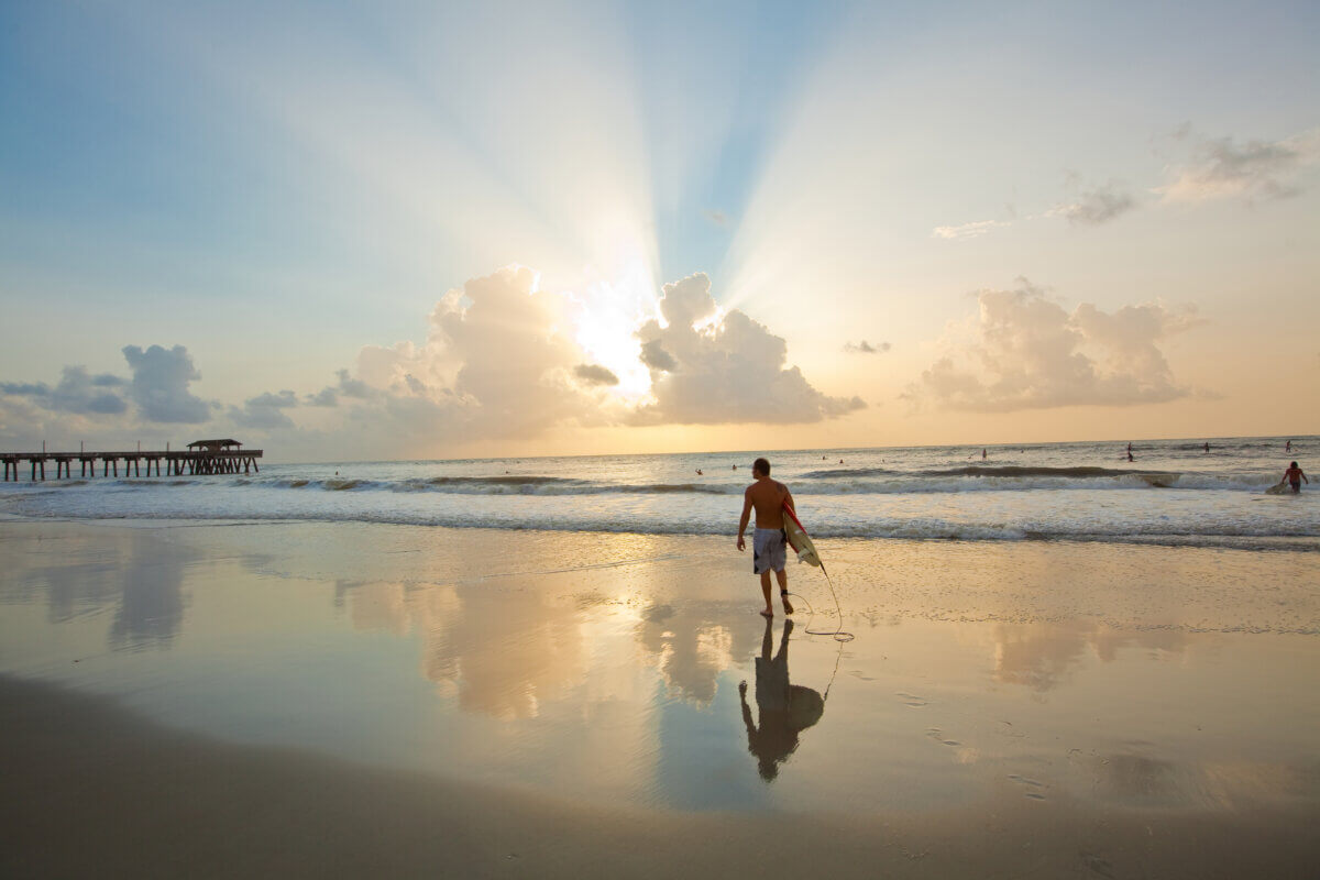 The Best Summer Weekend Getaways in the South - 3 A person walks along a sandy beach at sunset, holding a surfboard leash, with waves, clouds, and a pier in the background—capturing the best of summer weekend trips in the South.