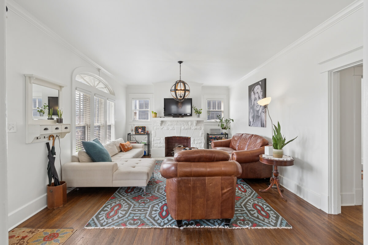 Bright living room at 1003 Mansfield Ave, Nashville TN, with white walls, wooden floor, a beige chaise sofa, two brown leather chairs, patterned rug, plants, and a fireplace with a TV and artwork above it.