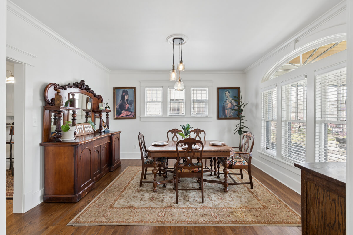 A traditional dining room at 1003 Mansfield Ave, Nashville TN, features a wooden table with six chairs, an ornate sideboard, portraits on the wall, large windows, and a patterned rug on a hardwood floor.