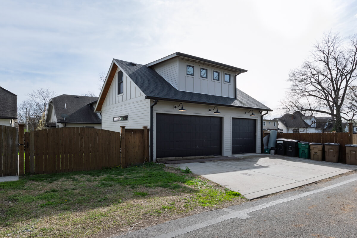 A two-story house at 1003 Mansfield Ave, Nashville, features three black garage doors, a sloped roof, a fenced yard, and multiple garbage bins lined up along the driveway.