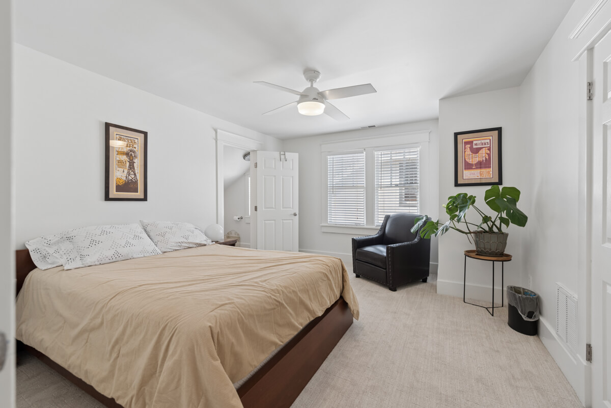 A bedroom at 1003 Mansfield Ave, Nashville TN features a large bed with tan bedding, an armchair by the window, framed art on white walls, a ceiling fan, and a potted plant.