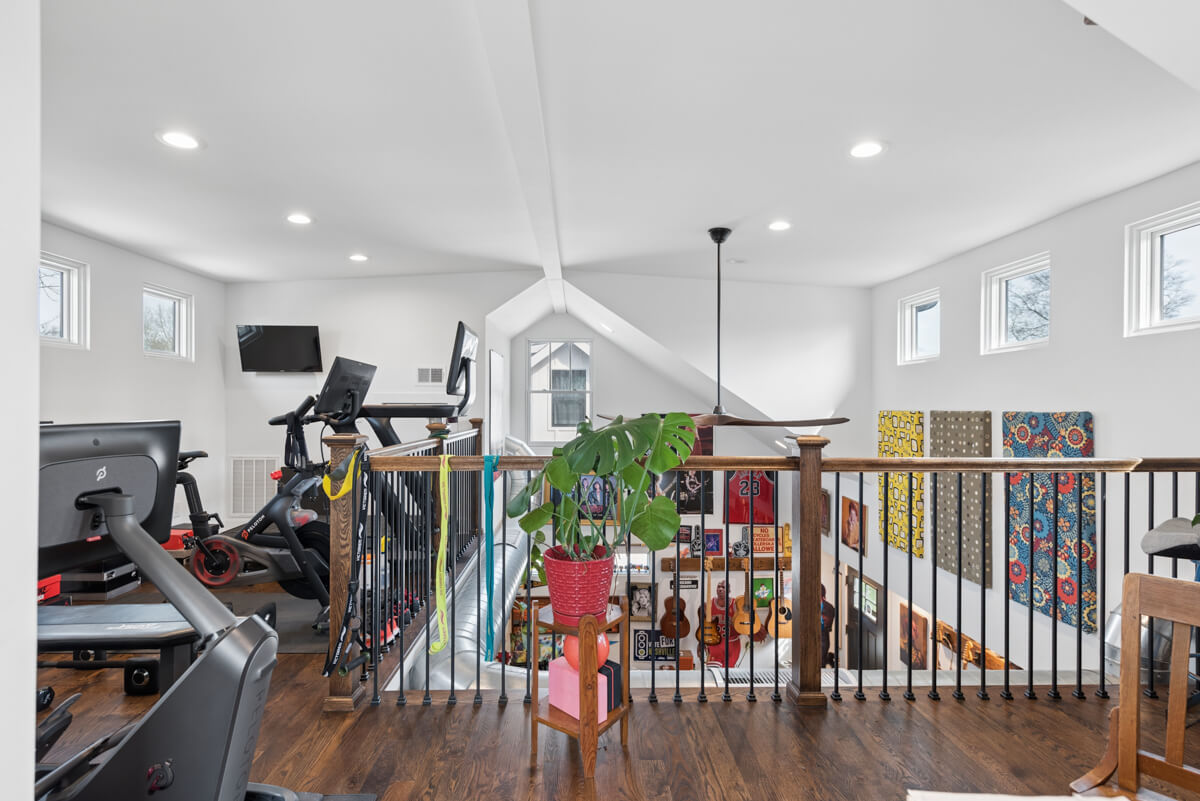 A home gym with exercise equipment, a potted plant on the railing, and wooden floors overlooks a colorful wall with guitars and art on the lower level—reflecting the vibrant spirit of Nashville at 1003 Mansfield Ave.