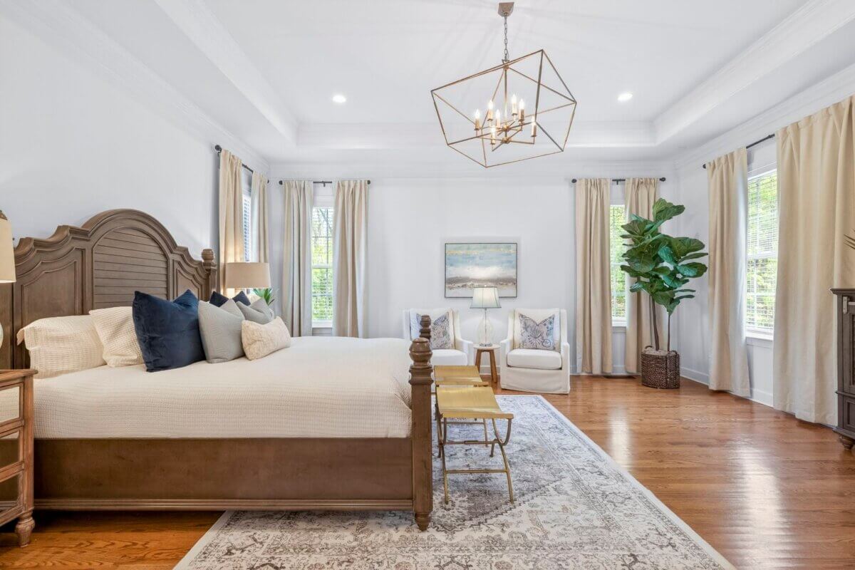 Spacious bedroom at 2708 Eglington, Franklin TN, features a large wooden bed, two benches, white armchairs, a potted plant, and neutral decor. Hardwood floors and large windows with beige curtains provide ample natural light.