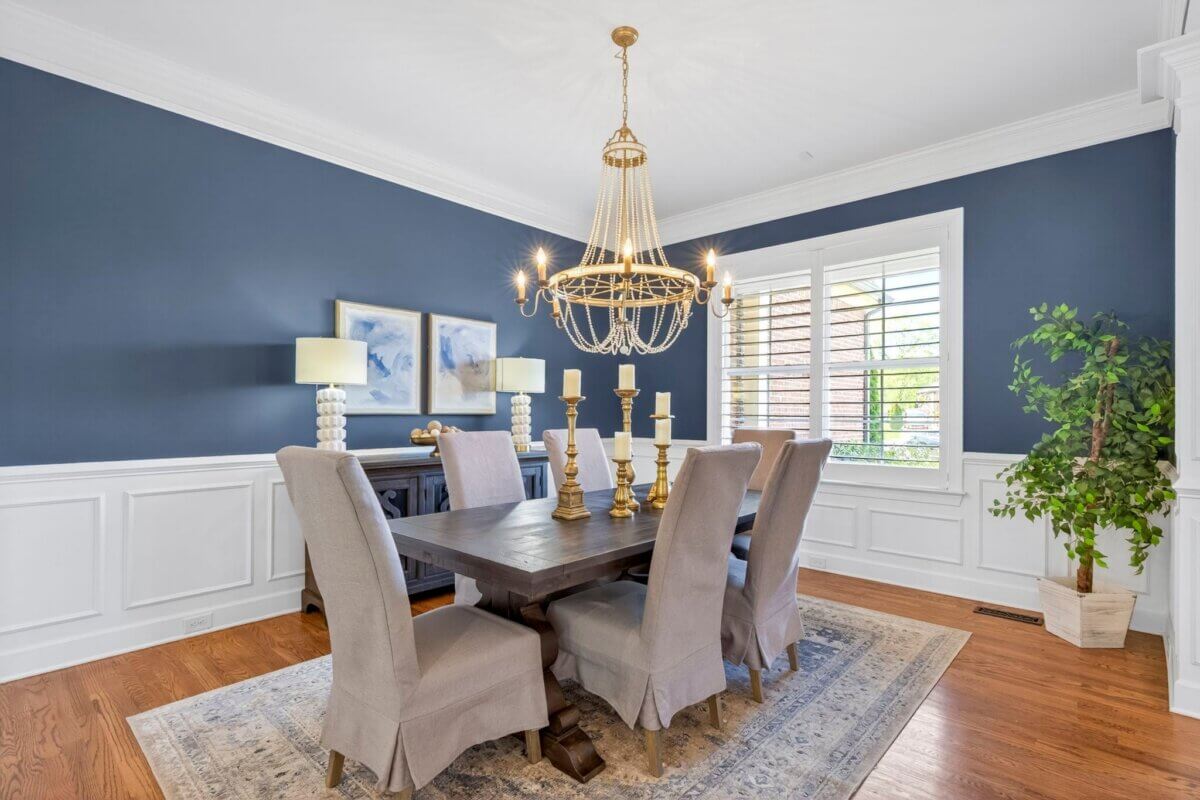 A stylish dining room in Eglington Terrace, Franklin TN, featuring a dark wood table, six upholstered chairs, a gold chandelier, blue walls with white trim, two lamps, framed art, a rug, and a potted plant. 2708 awaits your touch.