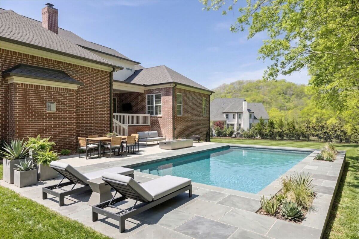 A backyard at 2708 Eglington Terrace features a rectangular swimming pool, lounge chairs, a dining table, and a brick house bordered by trees and lawn on a sunny Franklin TN day.