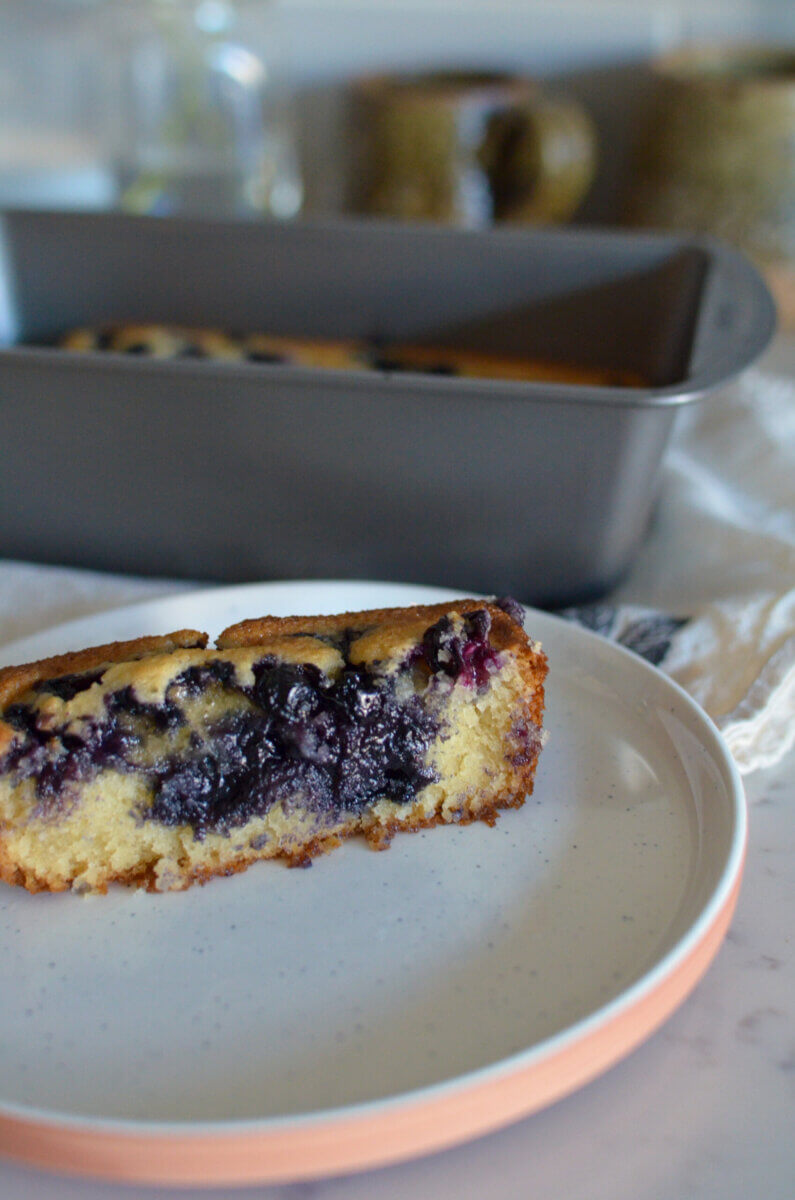 Two Summery Snack Cakes, Perfect for Picnics & Potlucks - 2 A slice of blueberry cake on a plate, with the rest of the cake still in a loaf pan in the background.