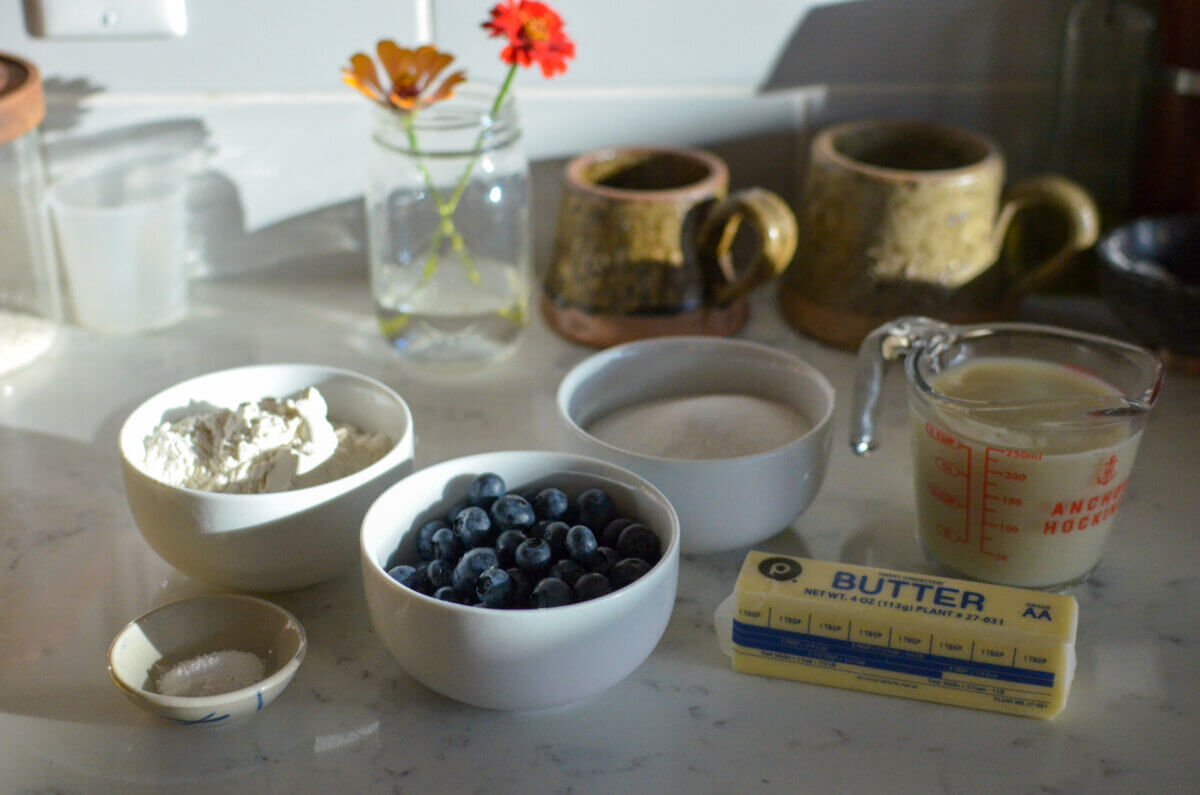 RECIPE: Easy Blueberry Buckle - 2 Ingredients for baking are laid out on a kitchen counter, including flour, blueberries, sugar, butter, milk, and salt, with mugs and flowers in the background.