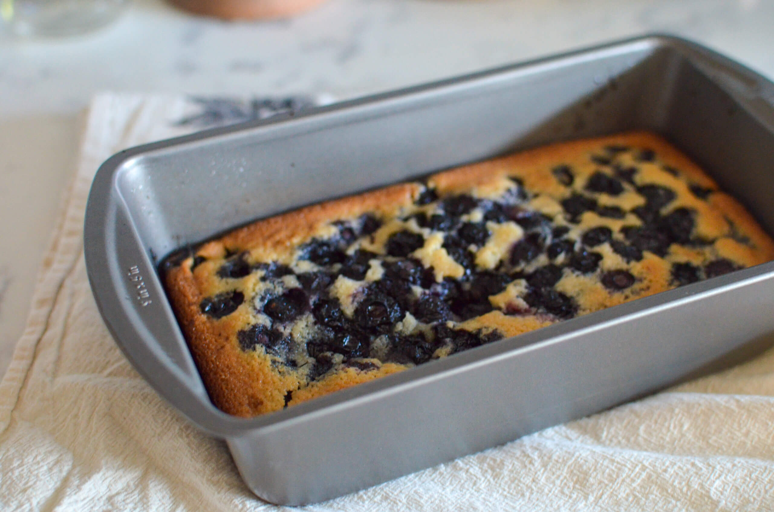 A rectangular metal baking pan holds a freshly baked blueberry cake resting on a light-colored cloth.