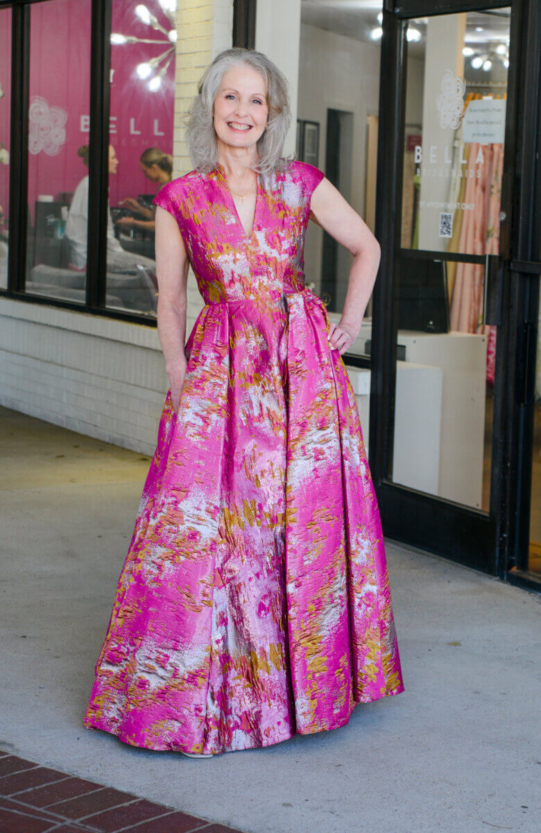 The Event-Season Trends Nashville Boutiques Are Loving Right Now - 7 A woman with gray hair stands outside a shop, All Dressed Up in a vibrant, floor-length pink and gold patterned dress, smiling at the camera.