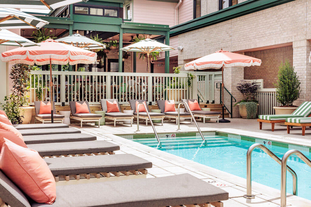 4 New Nashville Boutique Hotels That Should Be on Your Radar - 6 Outdoor pool area with gray lounge chairs, coral cushions, striped umbrellas, and surrounding plants in a sunlit courtyard.