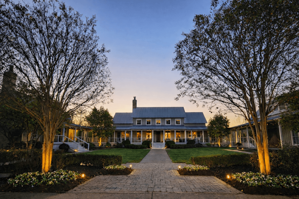 4 New Nashville Boutique Hotels That Should Be on Your Radar - 17 Large house with a metal roof, front porch, and columns, surrounded by manicured gardens and trees with exterior lights at dusk.