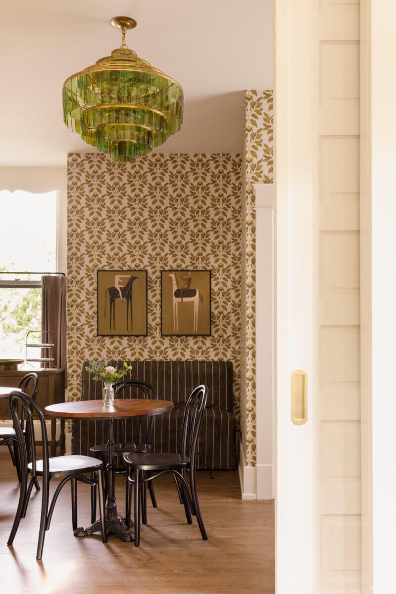 4 New Nashville Boutique Hotels That Should Be on Your Radar - 14 A dining area with a round wooden table, black chairs, floral wallpaper, framed art, a striped sofa, and a green glass chandelier. Light comes in from a nearby window.