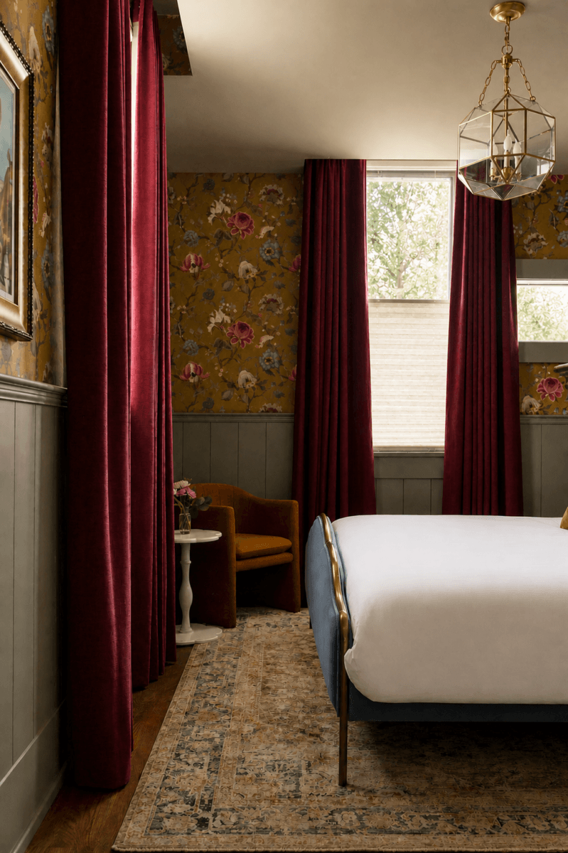 4 New Nashville Boutique Hotels That Should Be on Your Radar - 16 A bedroom with floral wallpaper, maroon curtains, an upholstered chair, a white side table, a brass bed frame, and a patterned rug.